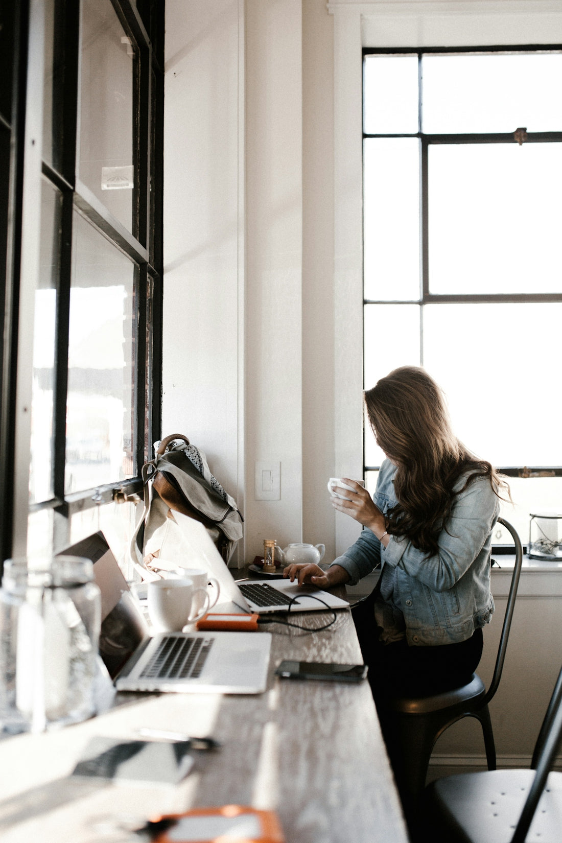 A woman in a denim jacket drinking coffee while working on a laptop